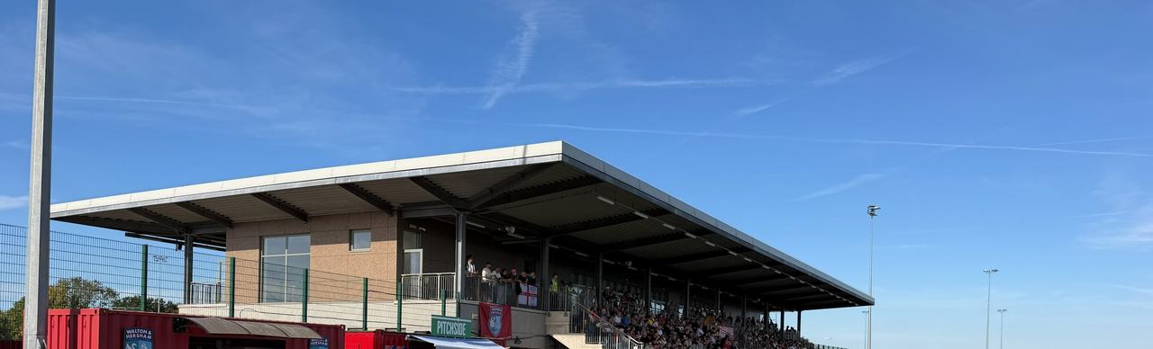 The covered stand at the Elmbridge Xcel Sports Hub on a sunny matchday, with spectators in the seats