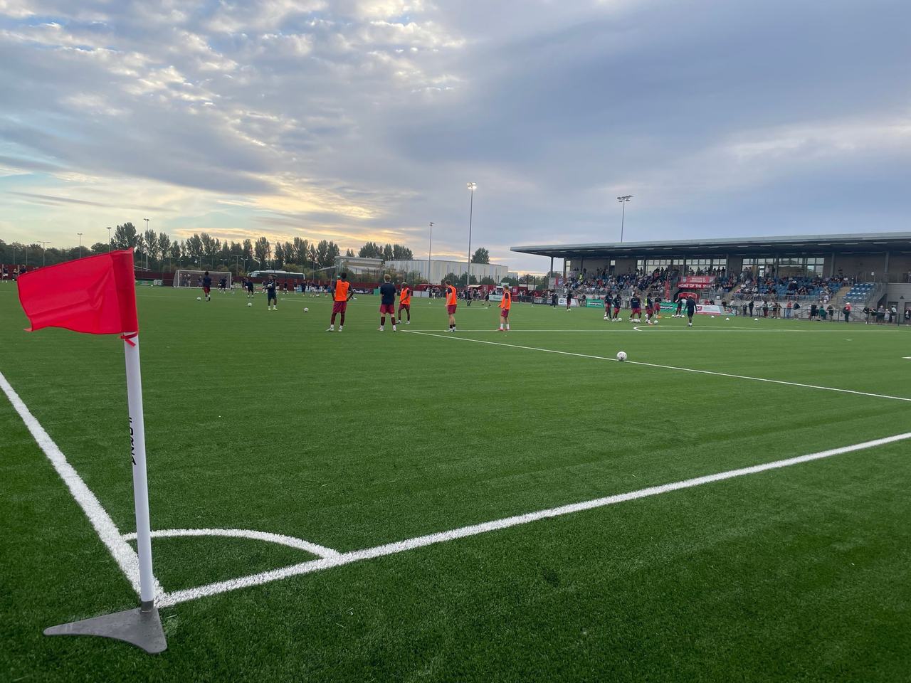 The pitch at the Elmbridge Xcel Sports Hub seen from a corner flag, with players warming up and the covered stand on the right