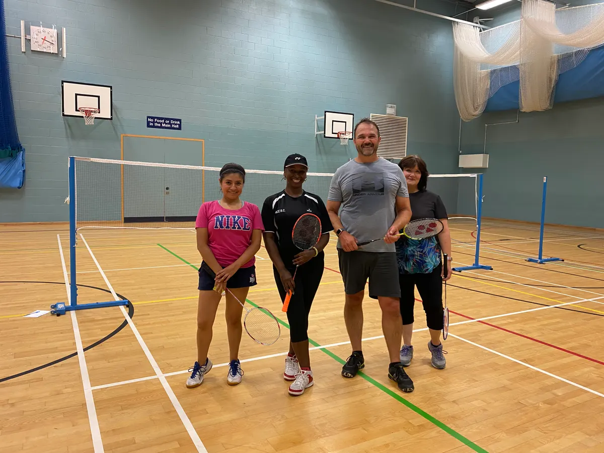 Four Ace Badminton Club players standing together on a court in a sports hall, holding rackets, one wearing a black club top printed ACE B.C.