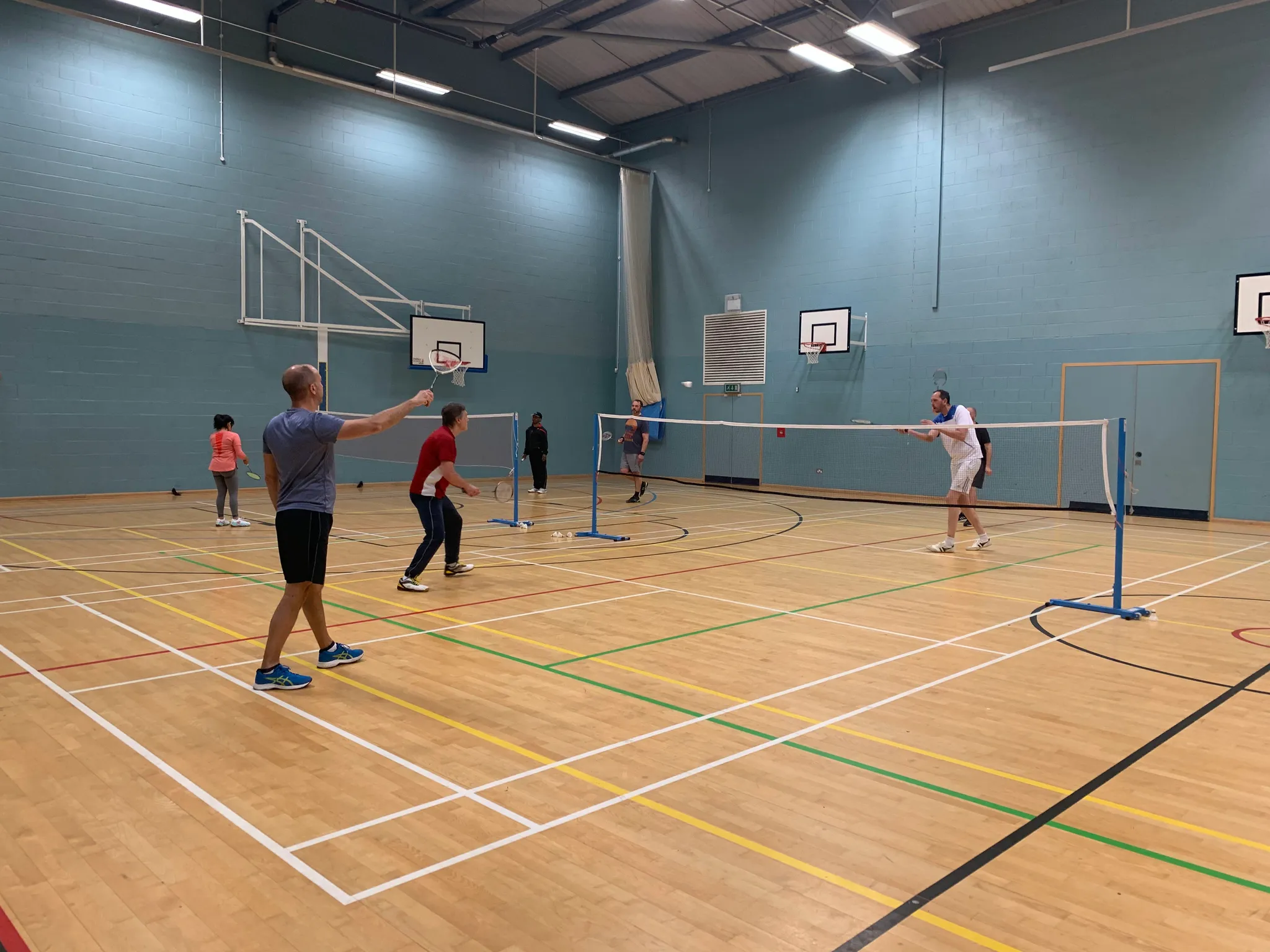 A sports hall with several badminton games under way, a player in a grey top in the foreground raising a racket towards the net