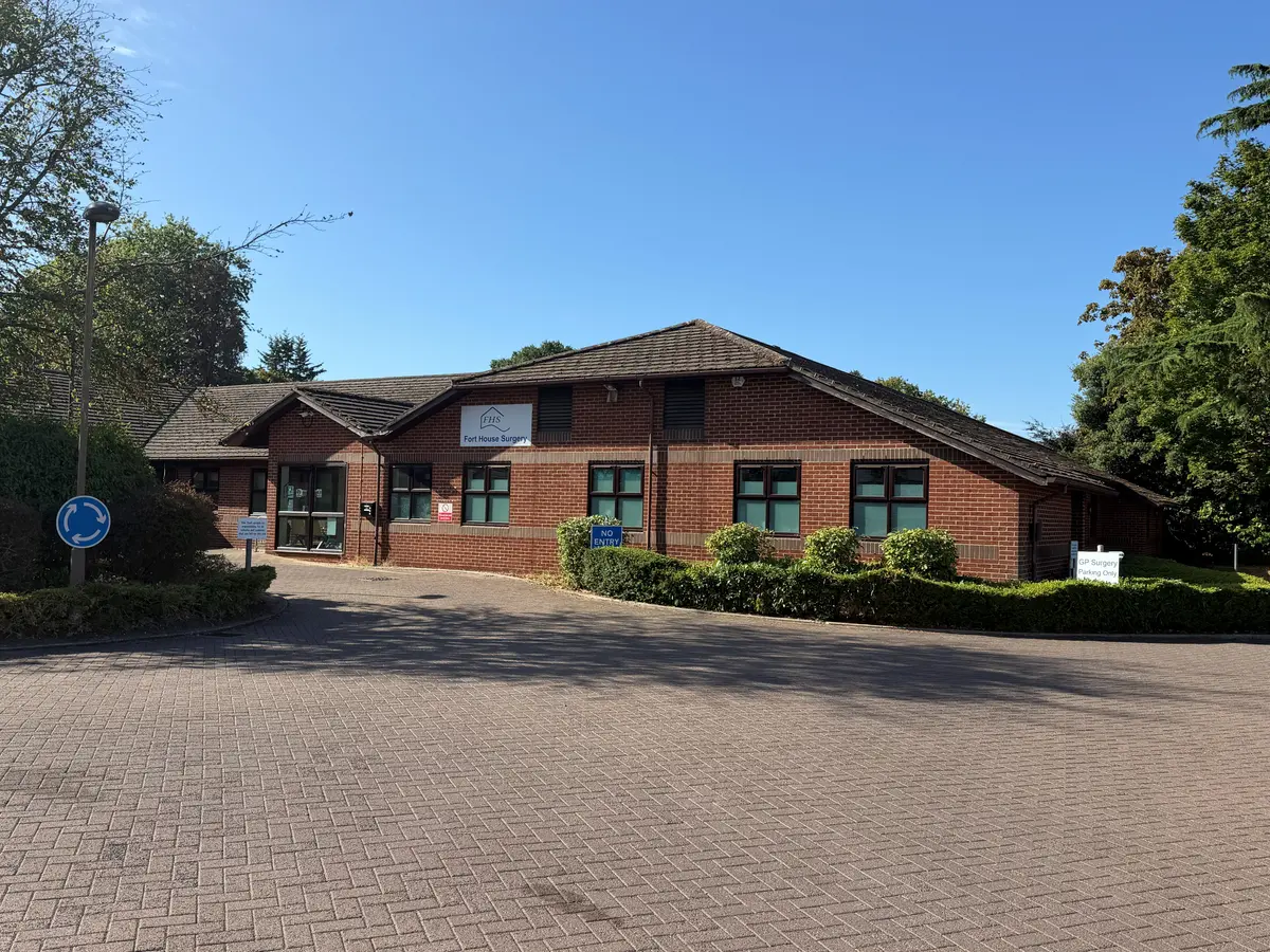 Fort House Surgery's single-storey red-brick wing at Walton Community Hospital, with the FHS Fort House Surgery sign above the windows and a GP Surgery Parking Only sign by the hedge on the right