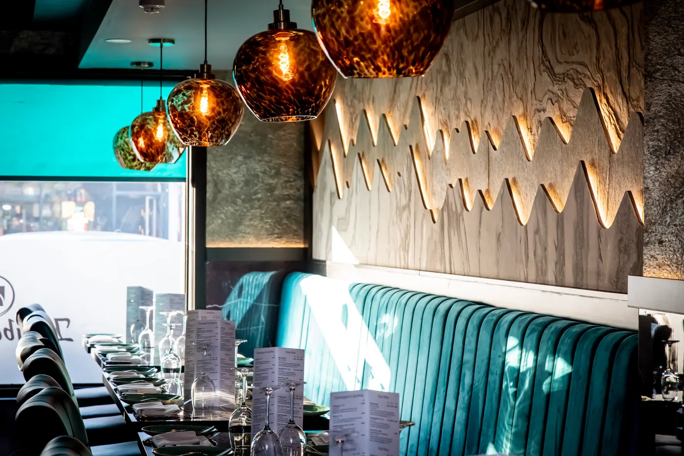 The dining room at Khyber Pass, with teal velvet banquette seating, copper glass pendant lights and a backlit carved wall panel