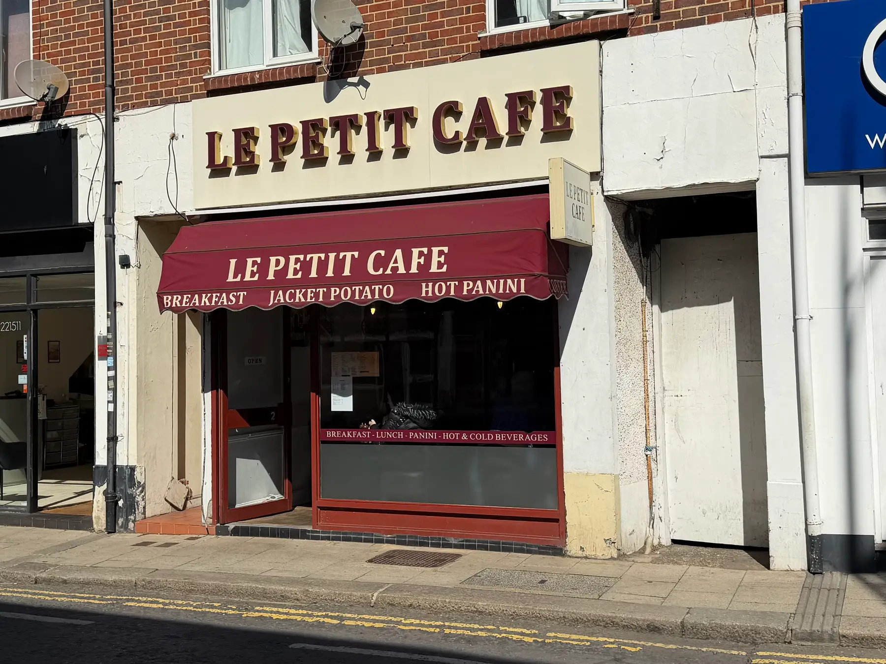 The shopfront of Le Petit Cafe on Bridge Street, Walton-on-Thames, its name in gold letters on a cream fascia above a burgundy awning reading Breakfast, Jacket Potato, Hot Panini