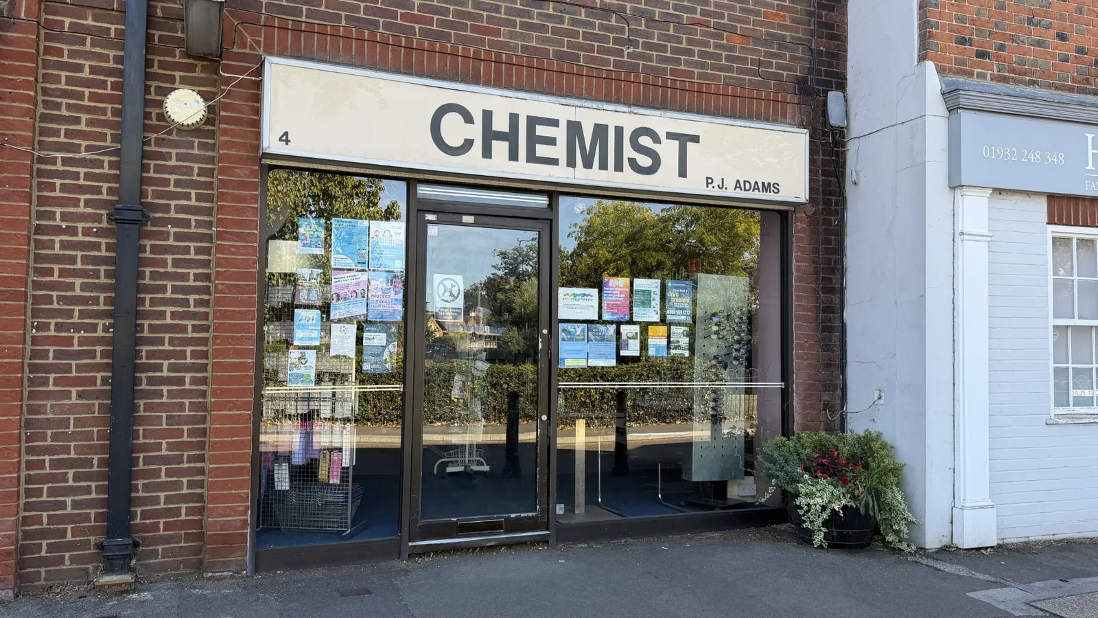 The shopfront of Philip J Adams Chemist at 4 Hersham Green, a glass door between two display windows under a cream fascia reading Chemist, P.J. Adams