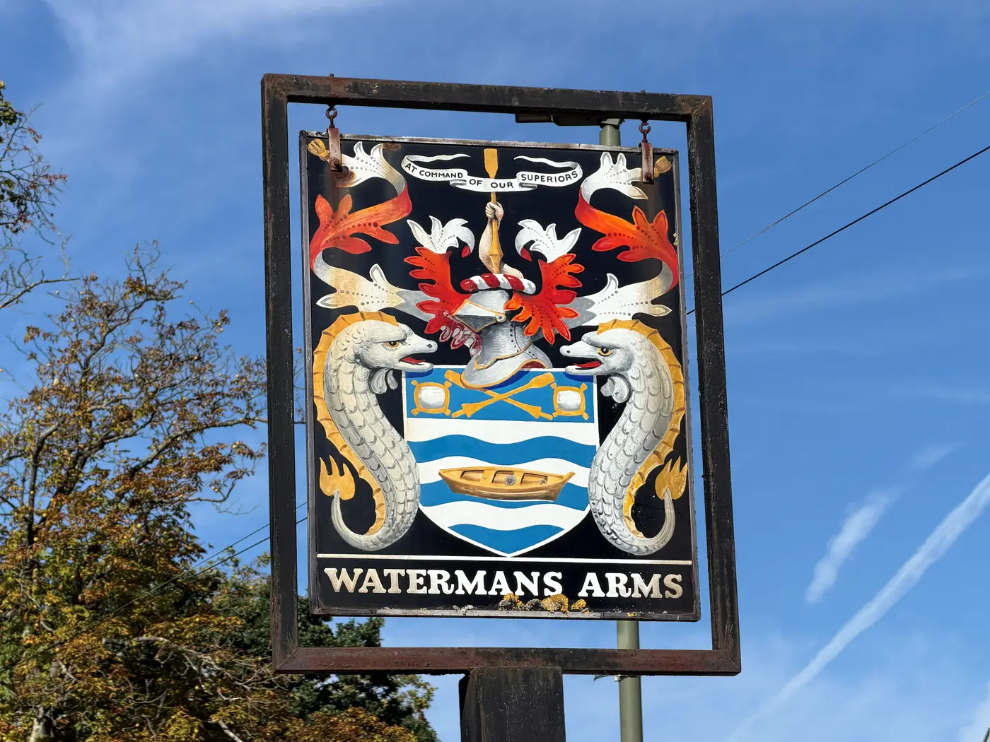 The hanging pub sign of the Watermans Arms, painted with a coat of arms bearing a boat on wavy blue and white bars, two silver sea-creature supporters and the motto At Command Of Our Superiors
