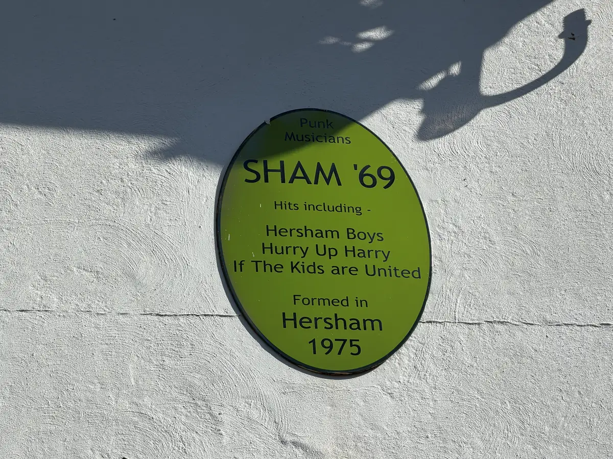 A green oval plaque on the pub's white wall reading Punk Musicians, Sham '69, hits including Hersham Boys, Hurry Up Harry, If The Kids are United, formed in Hersham 1975