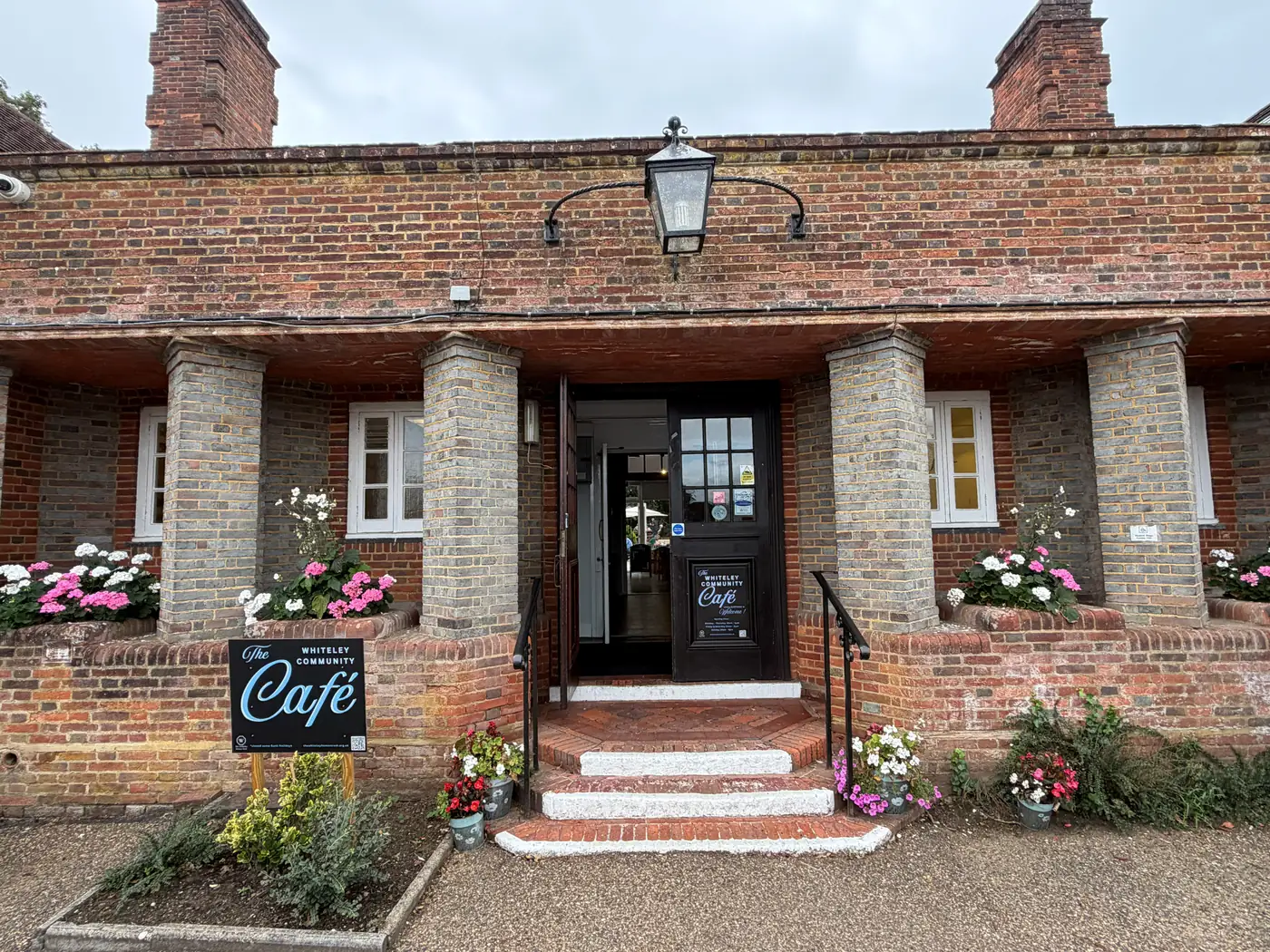 The brick-pillared porch and open front door of the Whiteley Community Café, with its signboard and flower-filled planters