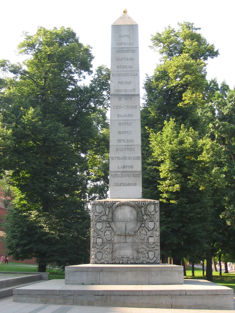 A tall grey stone obelisk standing among trees in a Moscow park, its shaft carrying nineteen surnames inscribed in Cyrillic, one to each block, from Marx at the top to Plekhanov at the foot, above a carved base