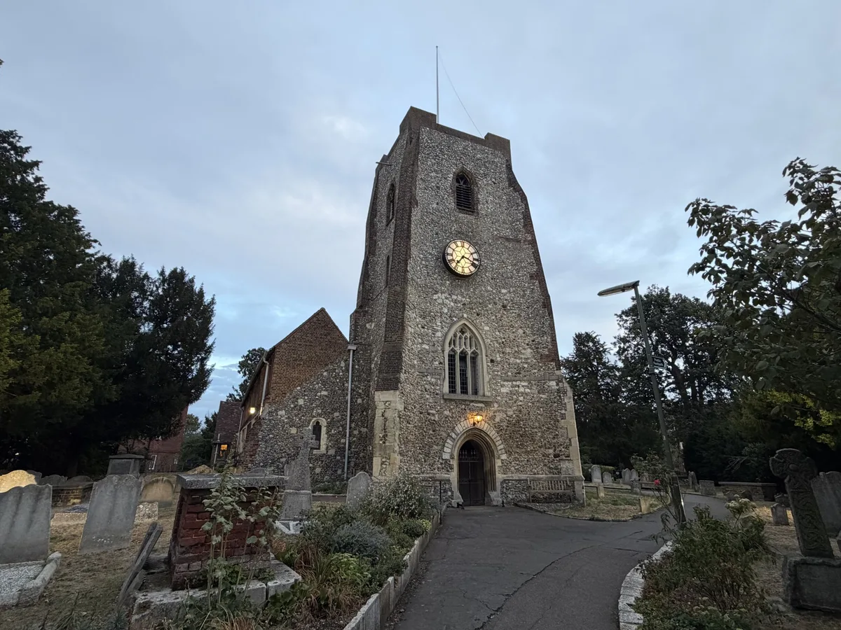 The flint and stone west tower of St Mary's Church at dusk, with a clock face, a traceried window and a lamp lit above the pointed arched doorway, approached by a path running between gravestones