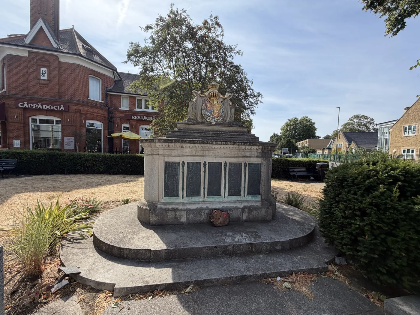 Front view of the Walton-on-Thames War Memorial with bronze name panels, stepped stone base and royal coat of arms, in a landscaped town-centre setting