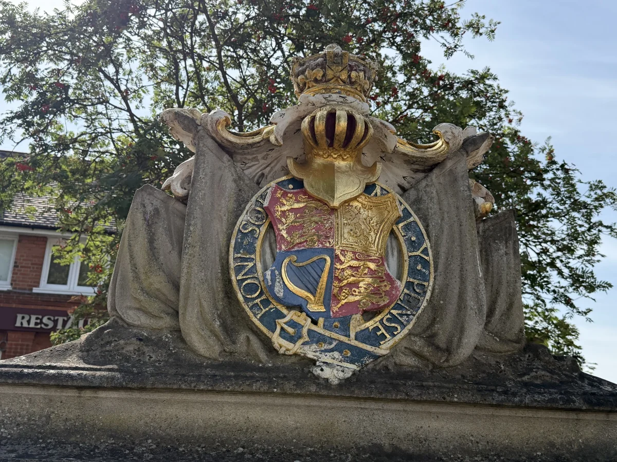 Close-up of the weathered royal coat of arms above the Walton-on-Thames War Memorial, with a crowned helmet, garter and draped stone flags