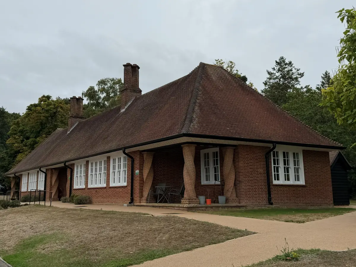 A long single-storey red-brick building with a steep hipped tiled roof, tall chimneys and twisted brick porch columns, set among trees