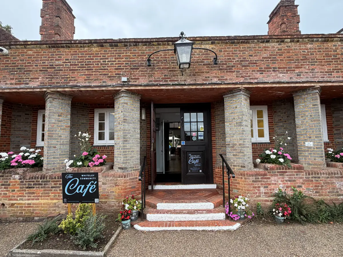 The brick-pillared porch and open front door of the Whiteley Community Café, with its signboard and flower-filled planters